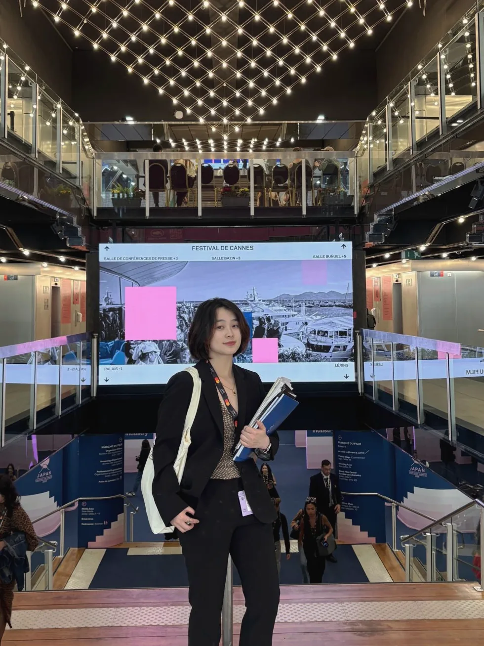 Yin Min in a black suit with a festival lanyard and a stack of folders, inside the Palais des Festivals.