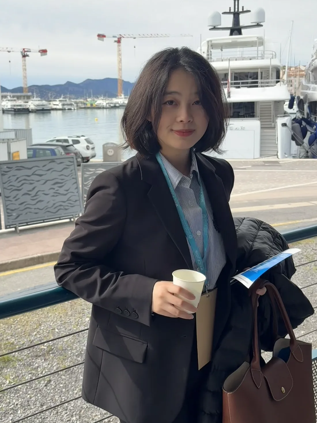 Yin Min in a black blazer with a conference lanyard and a coffee at the Cannes harbour, yachts behind her.