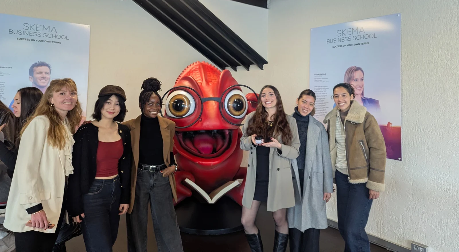 Seven people posing together in front of a large red chameleon statue at SKEMA Business School, one of them holding a small jar of skincare cream.